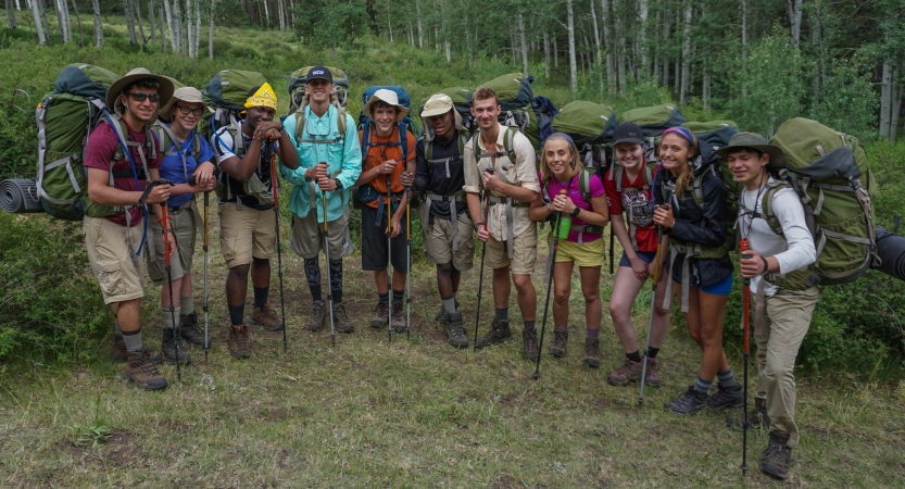 A group of students wearing backpacks smile for a photo in a green meadow 